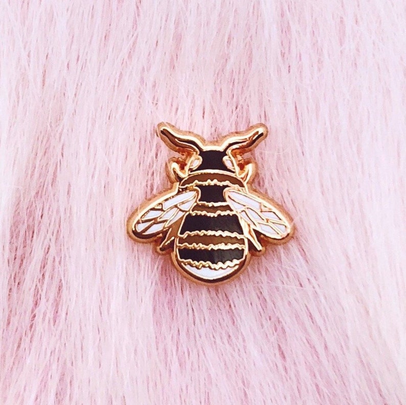 May include: A gold-toned bee pin with black and white stripes and white wings. The pin is on a pink fuzzy background.
