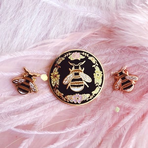 May include: Three gold-tone enamel pins featuring a bee design. The largest pin is a circle with a black background and a bee surrounded by flowers. The two smaller pins are simple bee designs.