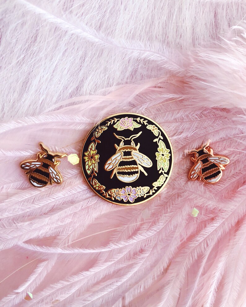 May include: Three gold-tone enamel pins featuring a bee design. The largest pin is a circle with a black background and a bee surrounded by flowers. The two smaller pins are simple bee designs.