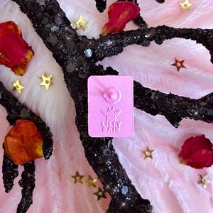 K&ouml;nnte beinhalten: Ein rosa Emaille-Anstecknadel mit dem Text "Lilly Baik" und einem floralen Design. Die Anstecknadel befindet sich vor einem Hintergrund aus rosa Federn, goldenen sternf&ouml;rmigen Konfetti und einem schwarzen, glitzerbedeckten Zweig mit roten Bl&uuml;tenbl&auml;ttern.