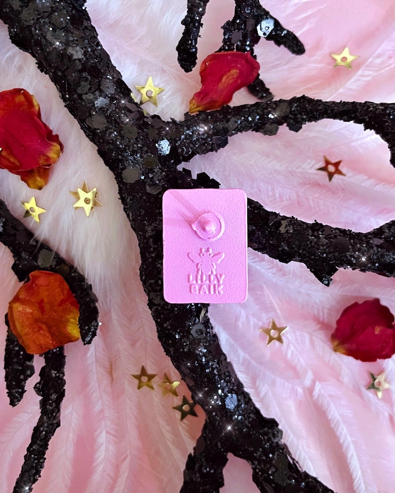 K&ouml;nnte beinhalten: Ein rosa Emaille-Anstecknadel mit dem Text "Lilly Baik" und einem floralen Design. Die Anstecknadel befindet sich vor einem Hintergrund aus rosa Federn, goldenen sternf&ouml;rmigen Konfetti und einem schwarzen, glitzerbedeckten Zweig mit roten Bl&uuml;tenbl&auml;ttern.