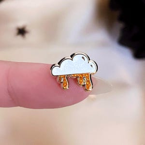 May include: A small, gold-coloured cloud-shaped pin with white enamel and dripping orange enamel details. The pin is a whimsical accessory, likely for clothing or bags.