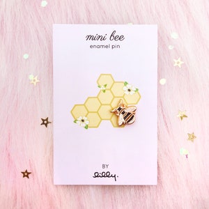 May include: A gold enamel pin featuring a cartoon bee on a honeycomb background. The pin says "mini bee enamel pin" and "by silly."