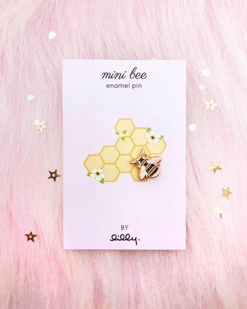 May include: A gold enamel pin featuring a cartoon bee on a honeycomb background. The pin says "mini bee enamel pin" and "by silly."