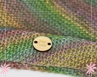 May include: Close-up of a hand-knitted scarf in shades of green, purple, and yellow. A small, round wooden tag with the text "Talk Dorky" is attached to the scarf. The knitted fabric has a textured appearance.