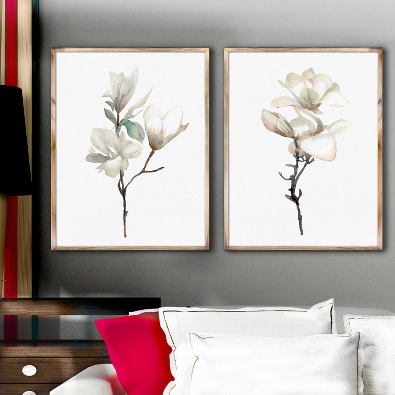Pode incluir: Duas pinturas de aquarela emolduradas de flores de magn&oacute;lia brancas sobre um fundo branco. As pinturas est&atilde;o em molduras douradas simples e est&atilde;o penduradas em uma parede cinza.