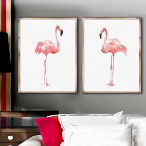 May include: Two framed watercolor paintings of pink flamingos on a white background. The flamingos are standing on one leg with their necks curved.