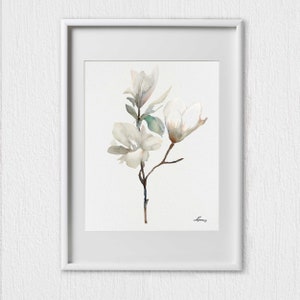 Pode incluir: Uma pintura em aquarela de um ramo de flor de magn&oacute;lia branca com folhas verdes. A pintura est&aacute; emoldurada em uma moldura branca.