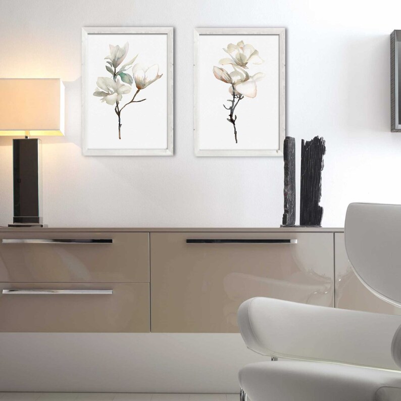 Pode incluir: Duas pinturas de aquarela emolduradas de flores de magn&oacute;lia brancas com folhas verdes sobre um fundo branco. As pinturas est&atilde;o penduradas em uma parede branca acima de uma c&ocirc;moda marrom claro com puxadores de gavetas prateados.