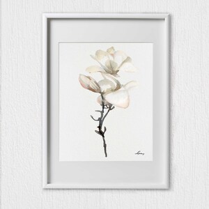 Pode incluir: Uma pintura em aquarela de uma flor de magn&oacute;lia branca com um caule e folhas delicados. A pintura est&aacute; emoldurada em uma moldura branca.