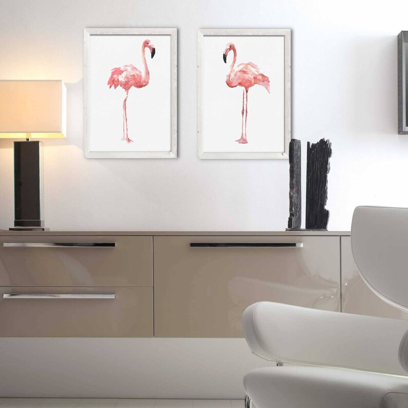 May include: Two framed watercolor paintings of pink flamingos, each flamingo is standing on one leg, the paintings are hanging on a white wall above a light brown cabinet with two drawers.