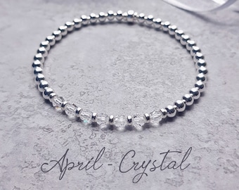May include: A silver beaded bracelet with clear crystal accents. The bracelet is on a gray background and the text "April - Crystal" is below the bracelet.