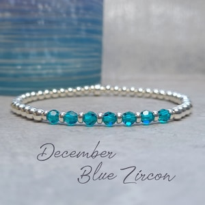 Op de afbeelding: Een zilveren armband met blauwe zirkoonstenen. Onder de armband staat de tekst "December Blue Zircon".