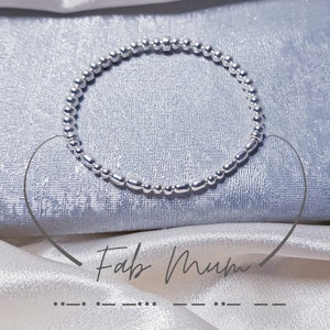 May include: Silver beaded bracelet with a silver bar charm. The bracelet is on a light blue fabric background. The text "Fab Mum" is written in cursive on the fabric.