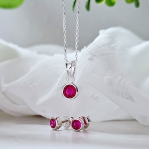 May include: A silver necklace with a pendant featuring a round, bright pink gemstone. Matching earrings with round, pink gemstones are also included.