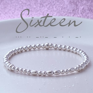 May include: A silver beaded bracelet with a silver charm. The bracelet is on a white surface and the word "Sixteen" is written above it.