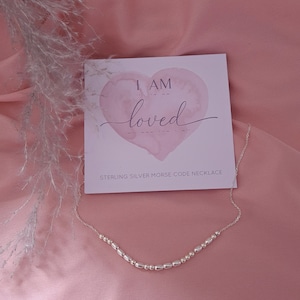 May include: Sterling silver necklace with small beads spelling out "I am loved" in Morse code. The necklace is displayed on a pink background with a card that reads "I am loved" in a pink watercolour heart.