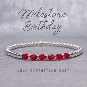 May include: A silver bracelet with red crystal beads. The bracelet has the text "Milestone Birthday" and "July Birthstone Ruby" on it.