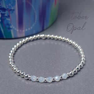 May include: A silver beaded bracelet with a row of five clear opal beads. The bracelet is on a grey surface and the text "October Opal" is written in black above the bracelet.