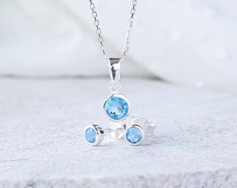 May include: A silver necklace with a blue gemstone pendant and matching earrings. The necklace has a delicate chain and the pendant is a round, faceted stone.
