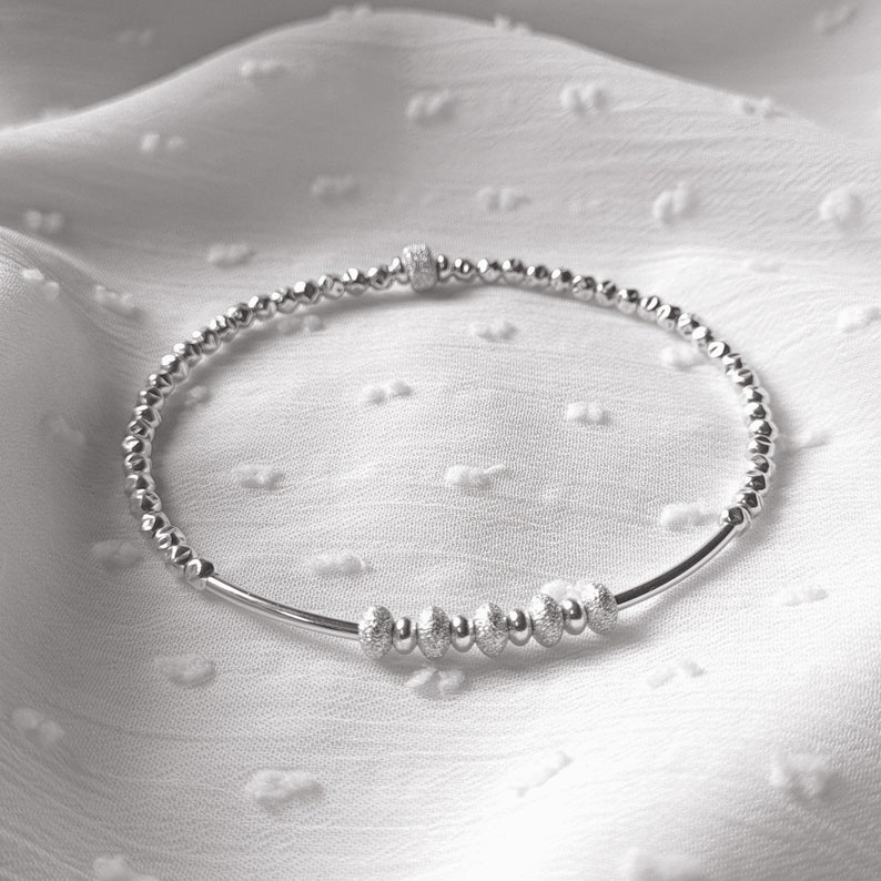 May include: A silver beaded bracelet with a small silver charm. The bracelet is made of tiny silver beads and has a single charm in the center.