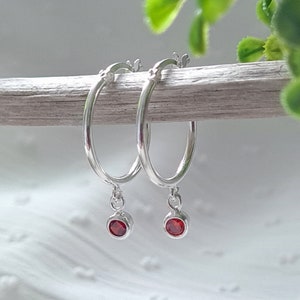 May include: A pair of silver hoop earrings with small red gemstone charms dangling from the bottom.