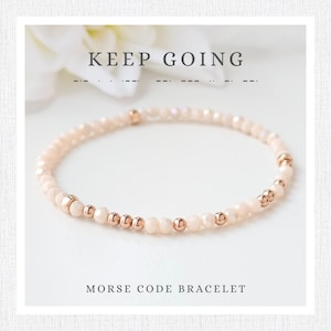 Op de afbeelding: Een delicate armband met perzik kleurige kralen en roségouden accenten. De tekst "KEEP GOING" staat erboven en "MORSE CODE BRACELET" eronder. De armband is ontworpen met kleine, ronde kralen en grotere, decoratieve kralen.