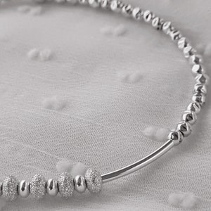May include: A silver bracelet with small, textured beads. The bracelet is made of a thin, silver band with a row of beads that are slightly larger and textured.