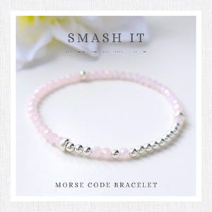 Può includere: Un braccialetto delicato con perline sfaccettate rosa chiaro alternate a piccole sfere metalliche argentate. Il testo SMASH IT e punti e linee del codice Morse sono visibili sopra, con la scritta MORSE CODE BRACELET riportata sotto.