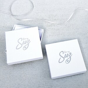 May include: Two white gift boxes with the text "One of a Suz" in a silver script font. The boxes are on a white textured surface.