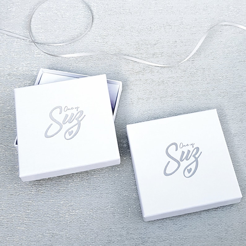 May include: Two white gift boxes with the text "One of a Suz" in a silver script font. The boxes are on a white textured surface.