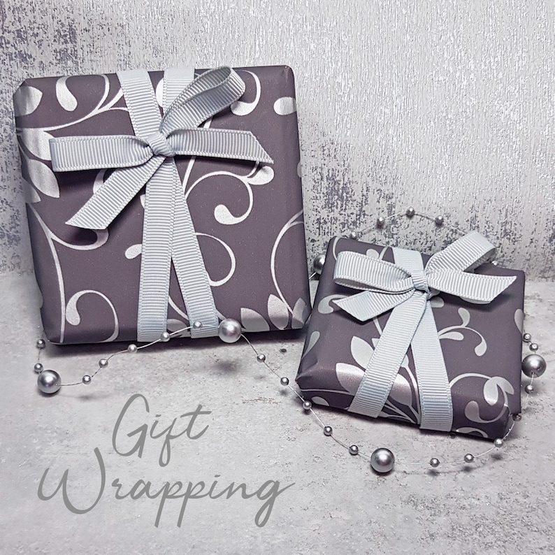 May include: Two silver gift boxes with white ribbon bows and a silver floral pattern. The boxes are on a white textured background with silver beads scattered around them. The text "Gift Wrapping" is written below the boxes.