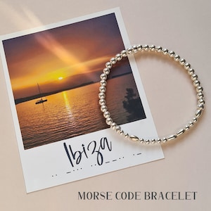 Peut inclure: Un bracelet de perles argentées avec un mélange de perles rondes et ovales. Le bracelet est présenté sur une carte postale avec une scène de coucher de soleil et le mot "Ibiza". Le texte "MORSE CODE BRACELET" est en dessous.
