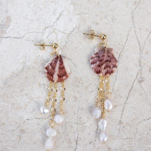 May include: A pair of gold-toned earrings featuring natural seashell pendants with a striped pattern in shades of brown and white. The earrings have delicate gold chains with dangling white pearl accents, creating a unique and elegant design.