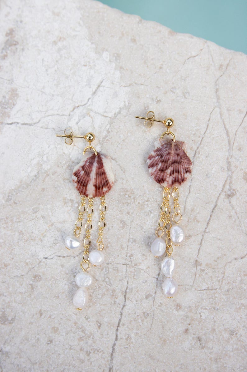 May include: A pair of gold-toned earrings featuring natural seashell pendants with a striped pattern in shades of brown and white. The earrings have delicate gold chains with dangling white pearl accents, creating a unique and elegant design.
