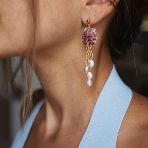 May include: Dangling earrings featuring a seashell design with a gold-colored post and chain. The seashell is a mix of pink and white hues, with several white pearls hanging from the chain. The earrings are a stylish accessory.