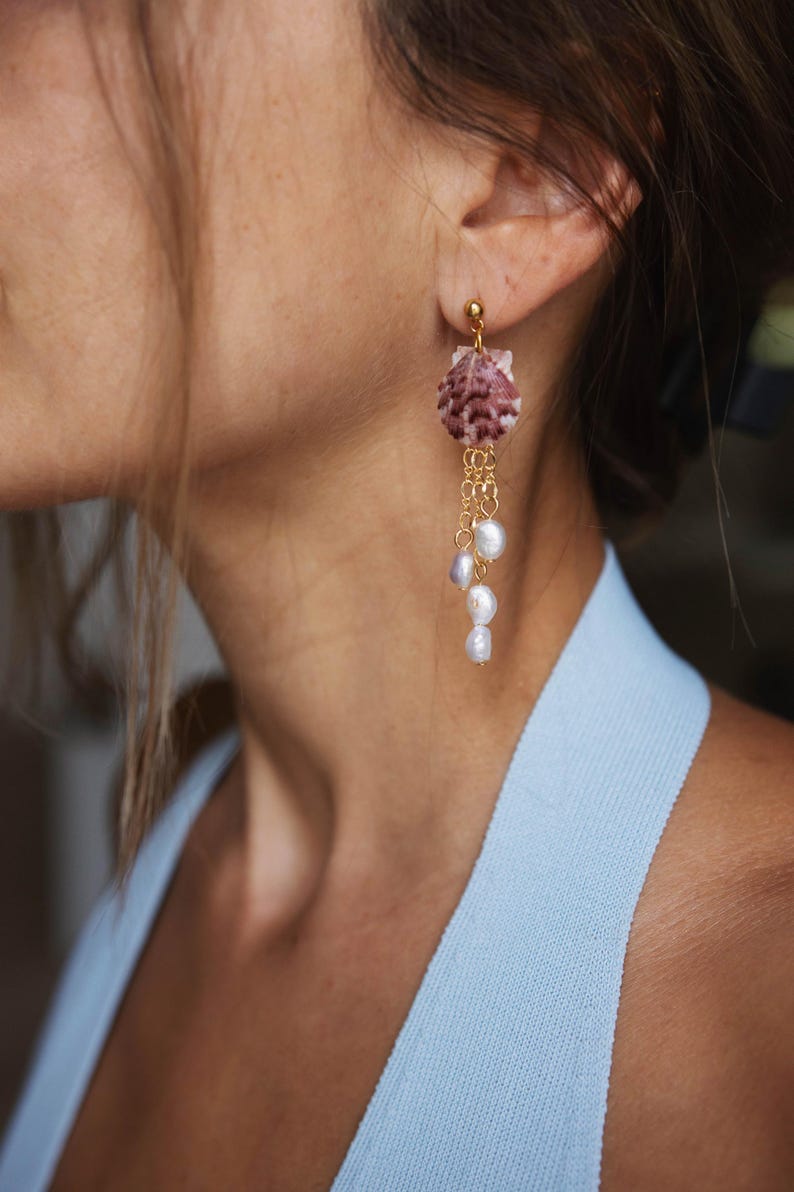 May include: Dangling earrings featuring a seashell design with a gold-colored post and chain. The seashell is a mix of pink and white hues, with several white pearls hanging from the chain. The earrings are a stylish accessory.