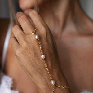 Könnte beinhalten: Ein goldfarbenes Handkettenarmband mit drei weißen Perlen. Die filigrane Kette verbindet sich mit einem Ring und umschließt das Handgelenk. Der Schmuck wird an einer Hand mit unscharfem Hintergrund getragen.