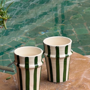 Puede incluir: Dos tazas de cerámica color crema con rayas verticales verdes. Las tazas tienen una parte superior acampanada y una superficie ligeramente texturizada. Están sobre una superficie marrón con un fondo de agua verde.