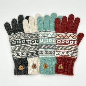 Może przedstawiać: Cztery pary dzianych rękawiczek w różnych kolorach: szary, biały, turkusowy i czerwony. Każda para ma biały, szary i czerwony geometryczny wzór. Każda rękawiczka ma małą brązową skórzaną metkę z logo alpaki.