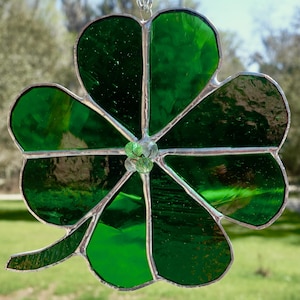 May include: A stained glass four-leaf clover suncatcher. The clover is made of various shades of green glass, with a silver-colored metal border. The suncatcher is hanging from a silver chain, perfect for St. Patrick's Day or as a lucky charm.