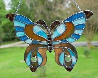 May include: A stained glass butterfly suncatcher with brown, blue, and white glass pieces. The butterfly is hanging from a silver chain.