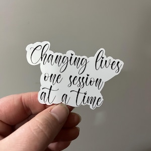 Puede incluir: Una pegatina blanca con texto negro en cursiva que dice "Changing lives one session at a time". La pegatina está sujeta por una mano sobre un fondo gris neutro. La pegatina tiene una forma única.
