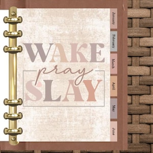 May include: A planner with a brown leather cover and gold metal rings. The planner's interior page has the words "WAKE pray SLAY" in a mix of neutral colors. Monthly tabs are visible on the right side, with the months January through June.