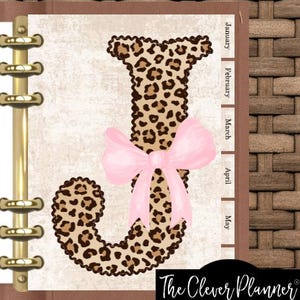May include: A planner page featuring a large letter J with a brown and black leopard print pattern, accented by a soft pink bow. The page includes side tabs labeled January, February, March, April, and May, and is held by a gold metal ring binder.