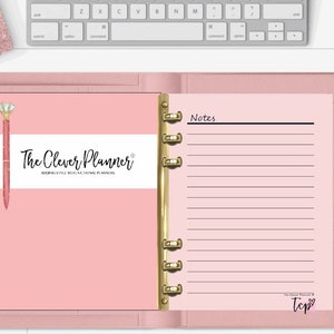 May include: A pink planner with gold accents, open to a lined page and a page with the text "The Clever Planner". A diamond-topped pen rests on the left page. A keyboard and accessories are in the background.