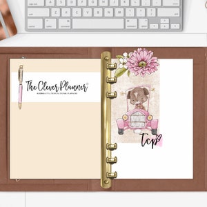 May include: A brown leather planner with a gold ring binder. The planner has a cream-colored page with the text "The Clever Planner" and a pink pen. The right page has a pink car with a brown dog driving and a pink flower.