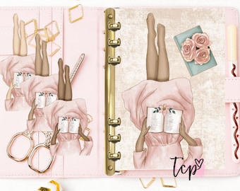 May include: A pink planner with gold rings and a decorative design featuring a woman reading a book. The design is repeated three times on the planner. The planner has a pink pen holder with a brown pen and a decorative brown and white design.