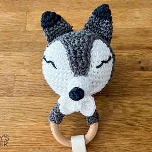 May include: A handmade crochet baby rattle shaped like a fox's head. The fox has a white face, gray and black ears, and closed eyes. It features a wooden ring and a white strap.