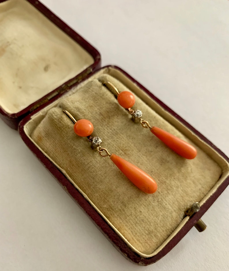 Pu&ograve; includere: Un paio di orecchini d'oro con dettagli in corallo e diamanti. Gli orecchini sono in una piccola scatola di velluto rosso.